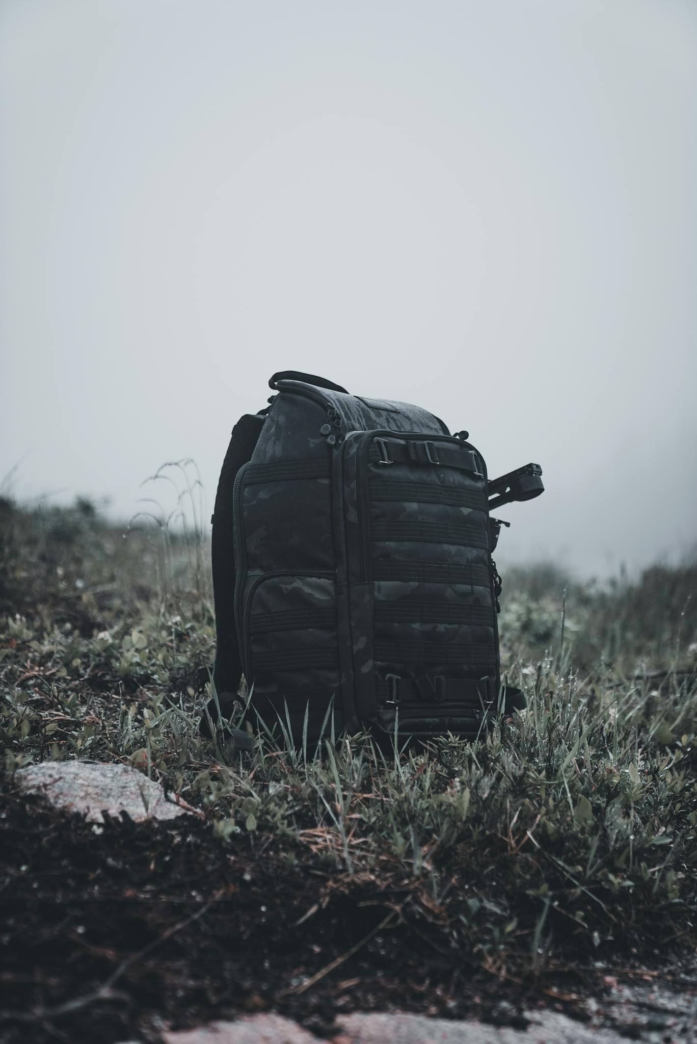 A rugged black backpack sits on grass in a misty outdoor environment, evoking a sense of adventure.