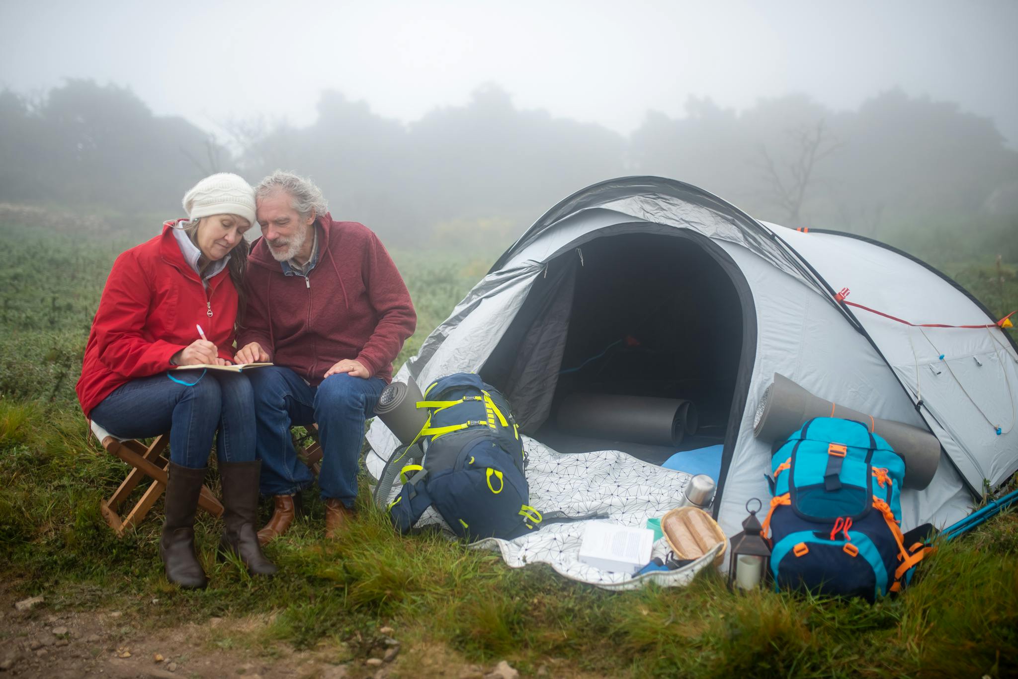 Senior couple enjoys quiet camping time in the Portuguese wilderness, seated by a tent on a foggy day.